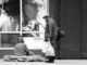 grayscale photo of man with shop bags walking past beggar siting on sidewalk